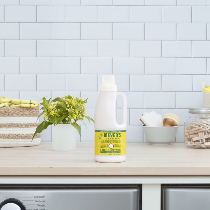 A bottle of Mrs. Meyers Fabric Softener, featuring essential oils, rests on a laundry rooms wooden table. Next to it are a potted plant and a basket of yellow towels against the clean backdrop of white tiled walls.