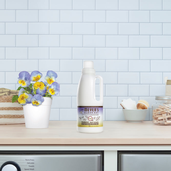 A bottle of Mrs. Meyers Fabric Softener infused with essential oils sits on a washing machine beside a pot of vibrant purple and yellow flowers, framed by a white tiled wall, alongside a bowl and glass jar of clothespins.