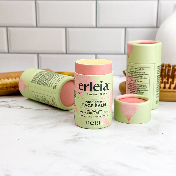 On the bathroom counter, Erleias skincare product tubes are displayed. Among them, the Face Balm - Acne Fighting stands upright, highlighting its vegan and cruelty-free formula. A wooden brush and white tile wall serve as the backdrop.