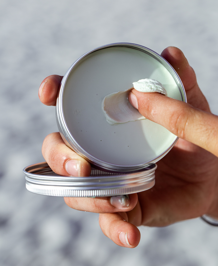 A hand holds an open recyclable tin of Raw Elements Face + Body - SPF 30, showcasing its smooth, creamy texture made from all-natural ingredients. Another finger scoops out some cream against a blurred outdoor background.