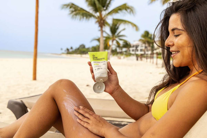 A woman in a yellow bikini applies Raw Elements Face + Body - SPF 30 natural sunscreen to her leg on a tropical beach, holding the tube of broad spectrum protection with palm trees and the ocean behind her. She smiles in the sunny, relaxing setting.