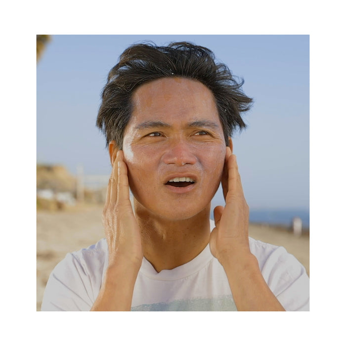 At the beach, under a clear blue sky with blurred scenery, a person in a white shirt applies Raw Elements Face + Body SPF 30 sunscreen for broad spectrum protection to their face using both hands.