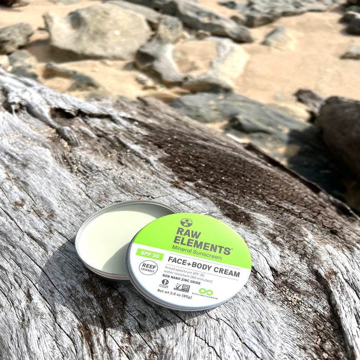 An open tin of Raw Elements Face + Body - SPF 30 sits on a weathered log by sand and rocks, showcasing its water resistance and broad spectrum UVA/UVB protection—ideal for sunny beach days.