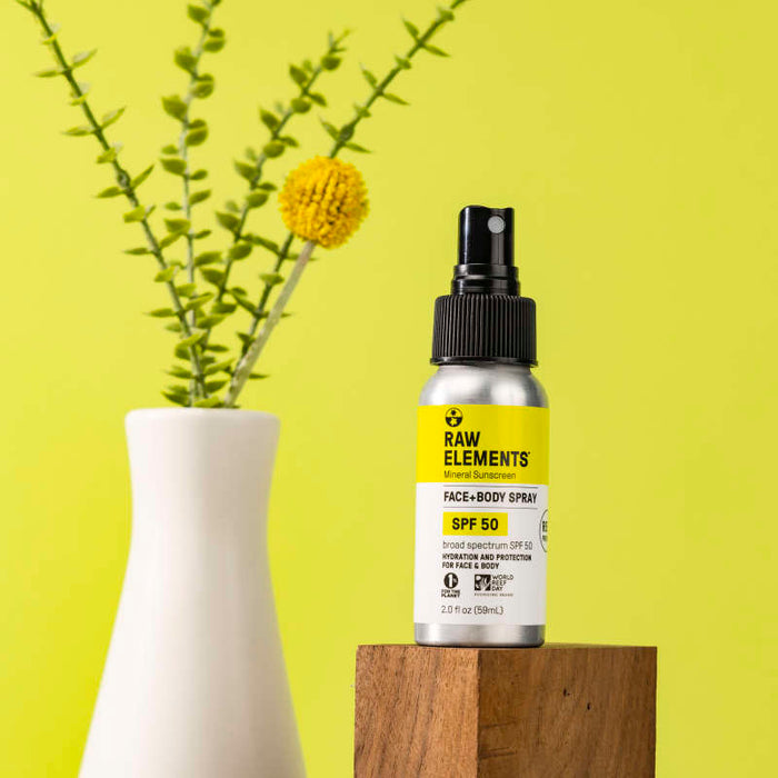 A metal spray bottle labeled Raw Elements Face & Body Spray - SPF 50 rests on a wooden block against a light yellow background, next to a white vase with green stems and a yellow spherical flower, showcasing its broad-spectrum protection.