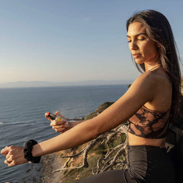 Standing on a cliffside, a person wearing a black crop top and leggings sprays Raw Elements Face & Body Spray - SPF 50 on their arm. They bask in broad spectrum protection, eyes closed as they gaze at the ocean while the setting sun casts a warm glow over the serene scene.