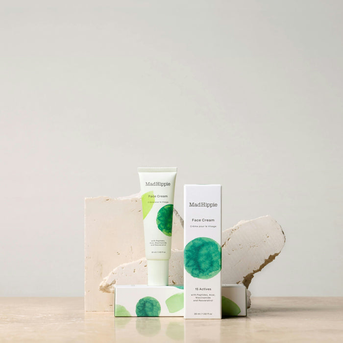 A tube and box of Mad Hippie Face Cream sit on a beige surface with stone accents, showcasing antioxidant-rich ingredients. The packaging has green and white design elements, set against a plain, light gray background.