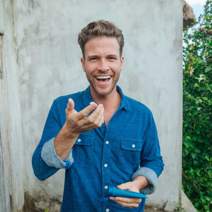 A smiling man in a blue button-up stands outdoors by a weathered wall, holding Oars + Alps Face + Eye Cream and gesturing, with greenery in the background.