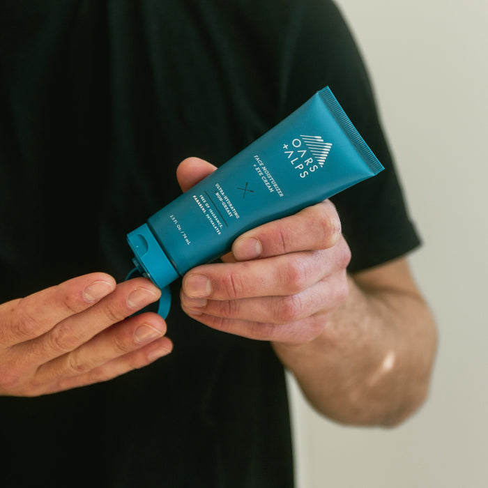 Someone in a black shirt holds a blue tube of Oars + Alps Face + Eye Cream, squeezing some onto their fingers.