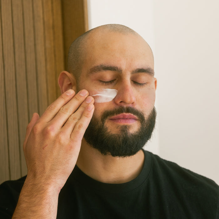 A bearded man with a shaved head, eyes closed, applies Oars + Alps Face + Eye Cream to his cheek with his fingers indoors by a wooden wall, wearing a black shirt.