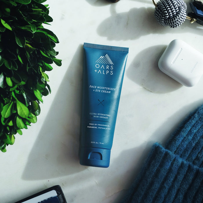 A blue tube of Oars + Alps Face + Eye Cream rests on a white surface, surrounded by a plant, AirPods case, and blue knit fabric. This ultra-hydrating cream helps support skin elasticity.