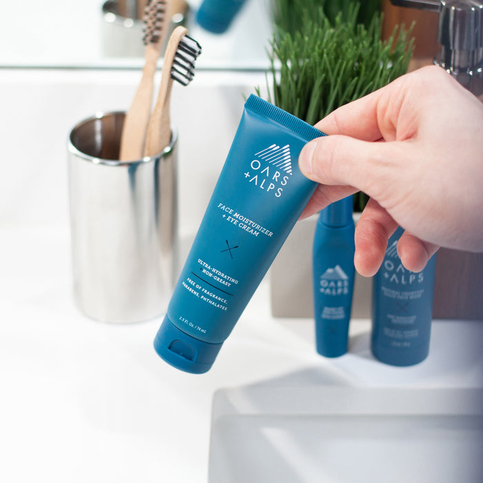 A hand holds a blue tube of Oars + Alps Face + Eye Cream over a bathroom sink, with toothbrushes and skincare products in the background, highlighting care for skin elasticity.