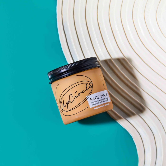 An UpCircle Face Mask with Kaolin Clay in a brown jar with a black lid rests on a cream surface against a teal background, showcasing its rejuvenating blend of cacao, coffee, and olive stones.