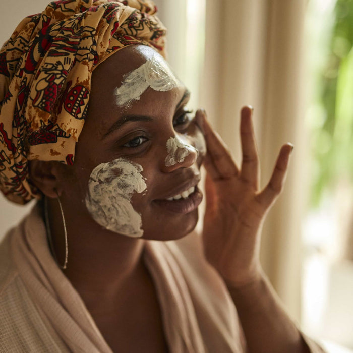 A woman in a vibrant headwrap and light robe applies UpCircles Face Mask with Kaolin Clay, smiling indoors with blurred greenery behind her.