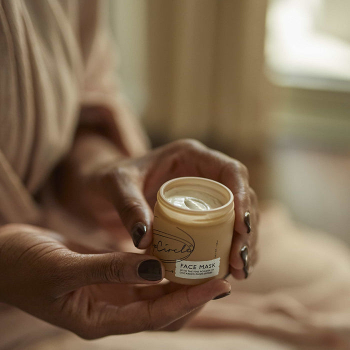 Close-up of hands holding an open beige jar labeled UpCircle containing a Face Mask with Kaolin Clay. Dark-painted nails contrast nicely, set against a softly blurred background that evokes a cozy indoor pampering atmosphere.