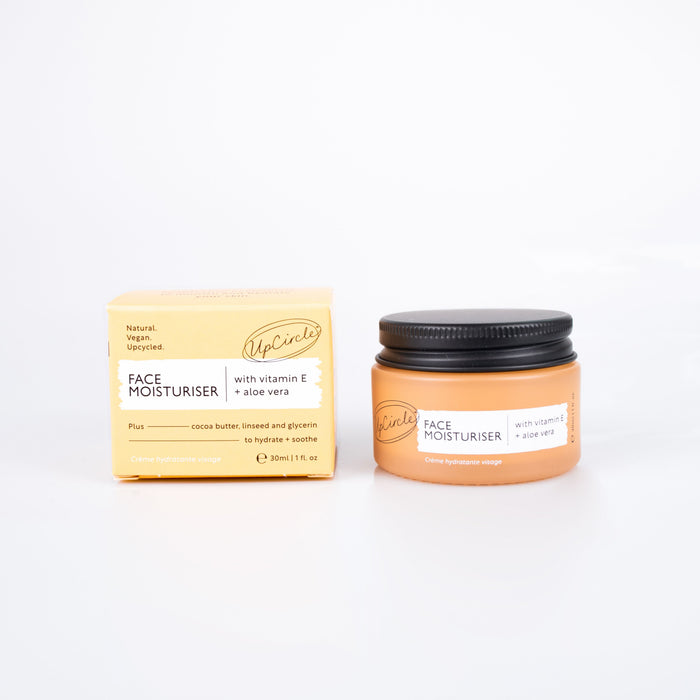 An UpCircle Face Moisturizer with Vitamin E and Aloe Vera, in an amber jar with a black lid, sits next to its box. The packaging highlights natural ingredients such as cocoa butter, glycerin, and vitamin E-rich argan powder that brighten and soothe the skin.
