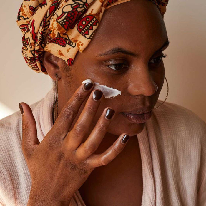 A person in a patterned head wrap and robe applies UpCircles Face Moisturizer with Vitamin E + Aloe Vera to their cheek. Their dark brown nails contrast with large hoop earrings, set against a softly lit backdrop.