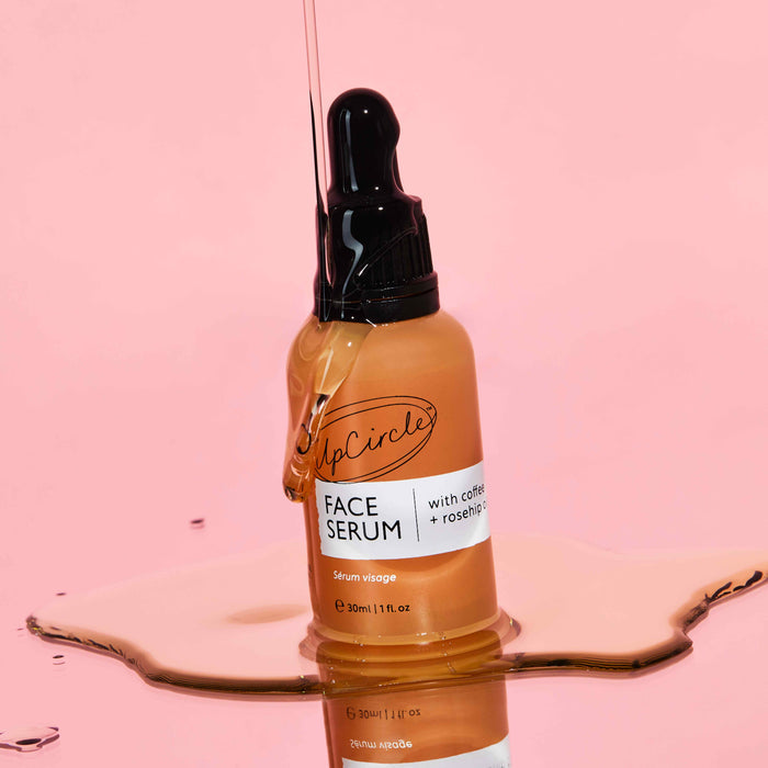 An UpCircle Face Oil bottle with a black dropper stands upright on a reflective surface against a pink background. The organic oil, featuring coffee, rosehip, and jojoba known for enhancing collagen production, spills gently pooling around the base.