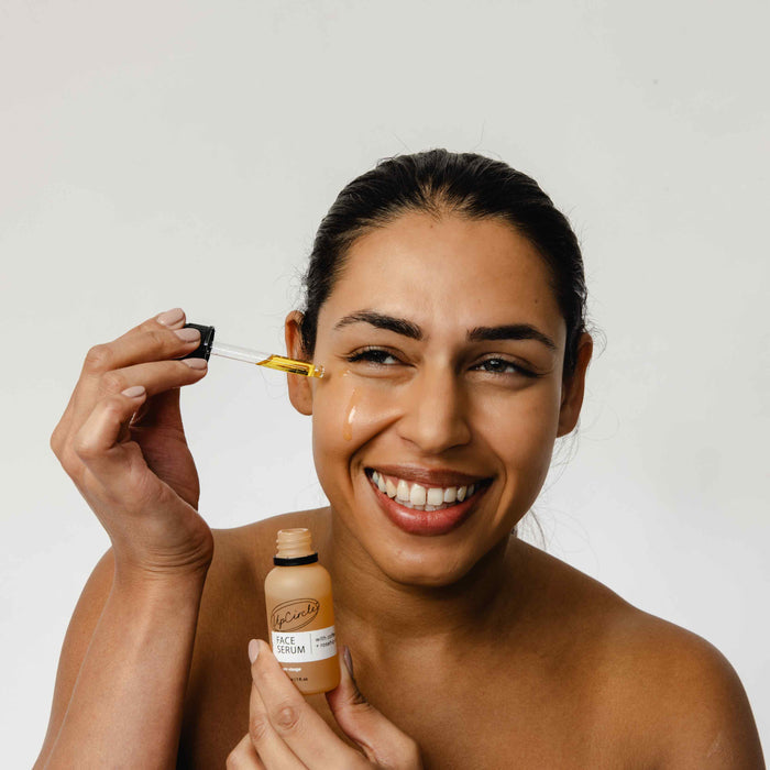 A woman smiles while applying UpCircles Face Oil with Coffee, Rosehip + Jojoba from a dropper onto her cheek. In her left hand, she holds a bottle labeled Face Oil. The plain white background accentuates the elegance of this skincare ritual.