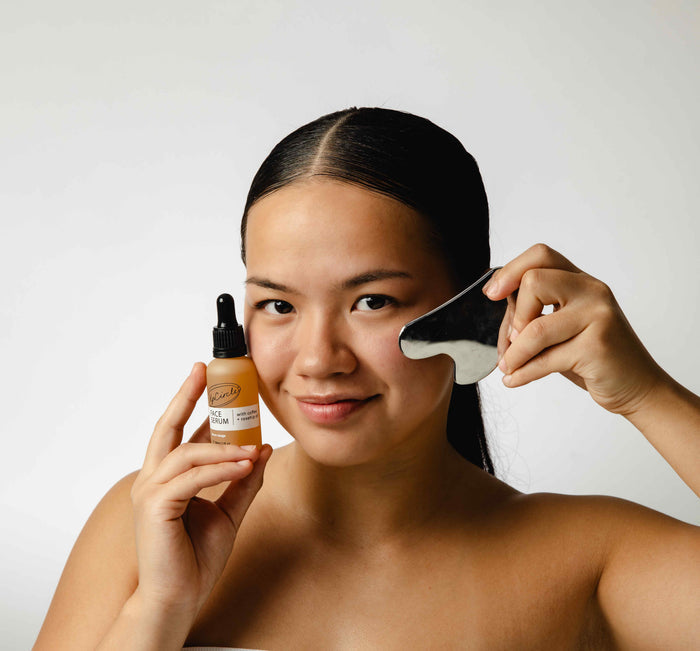 A woman with long dark hair holds a small skincare serum in one hand and UpCircles black facial massage tool infused with Face Oil with Coffee, Rosehip + Jojoba in the other. She applies it to her cheek against a plain white background, boosting collagen production smoothly.