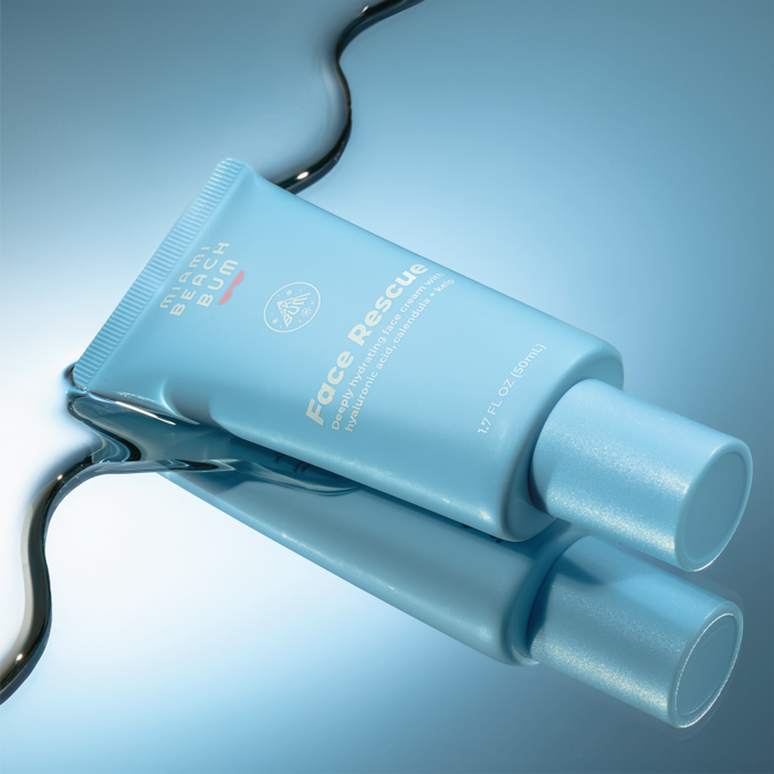 A light blue tube of Miami Beach Bums Face Rescue Cream rests on a glossy surface, reflecting its image like a mirror. White text highlights its promise to protect your moisture barrier.