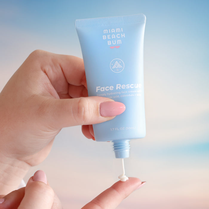 Someone holds a tube of Miami Beach Bum Face Rescue Cream, enriched with hyaluronic acid, dispensing a small amount onto their fingertip against a serene backdrop of pastel gradients.