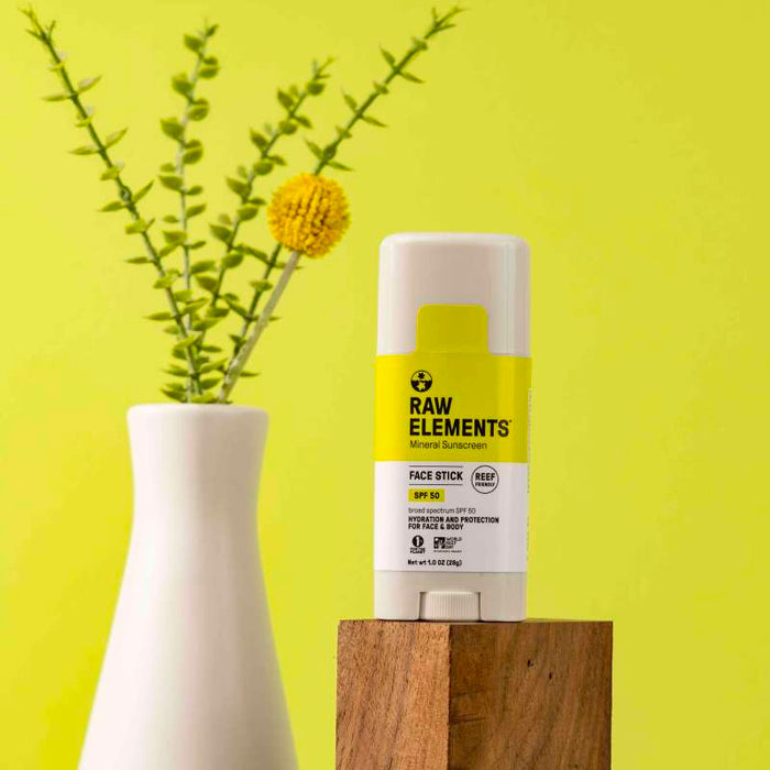 The Raw Elements Face Stick - SPF 50 Mineral Sunscreen is displayed on a wooden block against a bright yellow backdrop, offering broad spectrum protection. It is framed by a white vase with vibrant green and yellow plants.