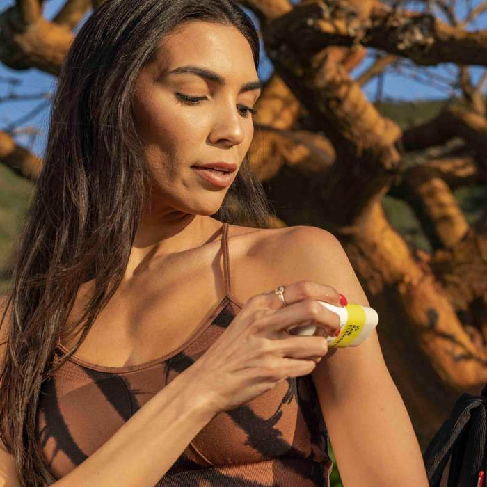 A woman outdoors applies Raw Elements Face Stick - SPF 50 to her shoulder. Shes in a brown tank top with a tree and blue sky in the backdrop.