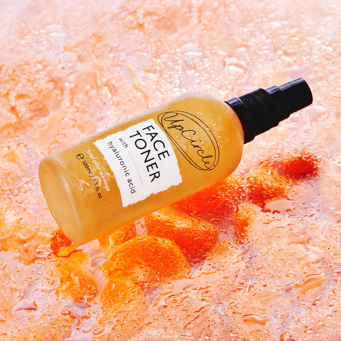 A bottle of UpCircles Face Toner with Hyaluronic Acid, featuring a spray cap, sits on an orange-toned surface with water droplets around it, enhancing its fresh appeal—ideal for any natural skincare routine.