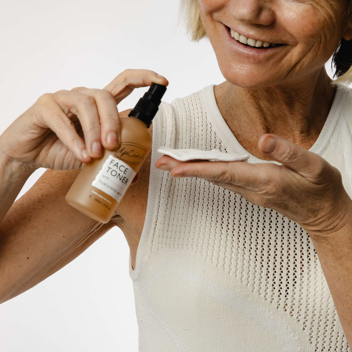 A person in a white sleeveless top smiles while applying hydrating toner onto a cotton pad. They hold an UpCircle bottle labeled Face Toner with Hyaluronic Acid featuring natural skincare ingredients in their left hand, against a plain white background.