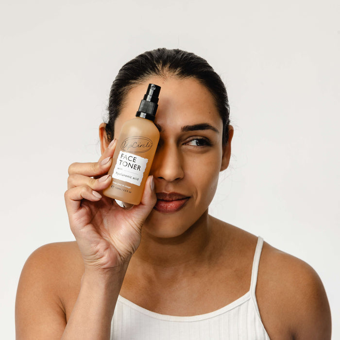 A person with tied-back hair holds a bottle of Face Toner with Hyaluronic Acid by UpCircle next to their face. Wearing a white tank top against a plain background, they embody the essence of natural skincare.