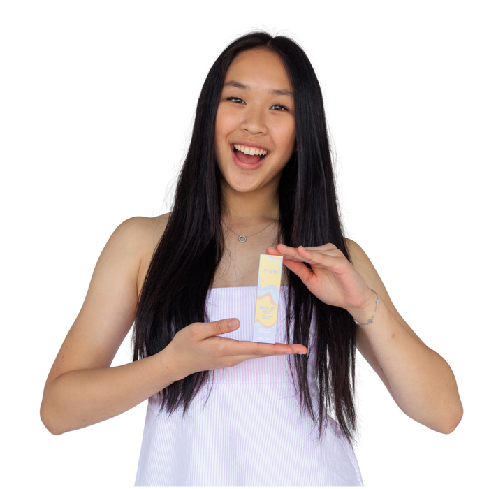 A smiling woman with long black hair holds a small box of Face Up To It Clarifying Dry Oil by evre. in yellow and white packaging, while wearing a sleeveless white top.