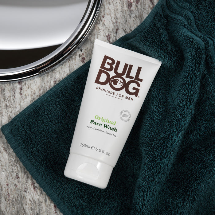 A tube of Bulldogs Face Wash featuring aloe, camelina, and green tea sits on a dark green towel. The cruelty-free skincare item is partially reflected in an adjacent round mirror.