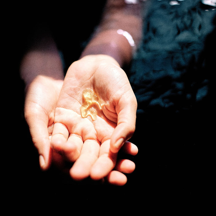 A close-up of an open hand holding a small, bright object like a gold leaf against a dark background. The persons torso, in deep blue or green fabric, is subtly visible. Light highlights the hand and object, evocative of discovering Thrive Natural Cares Face Washs precious essence.