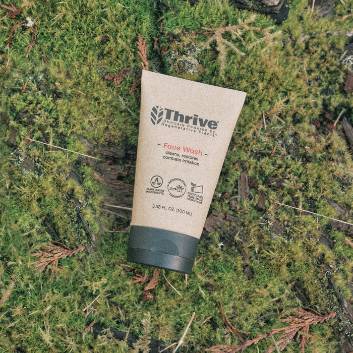 A tube of Thrive Natural Care Face Wash is set on moss and twigs, emphasizing its natural formula for cleansing, moisturizing, and soothing irritation. Eco-friendly symbols adorn the 3.38 fl oz (100 mL) packaging, showcasing its regenerative plant ingredients.