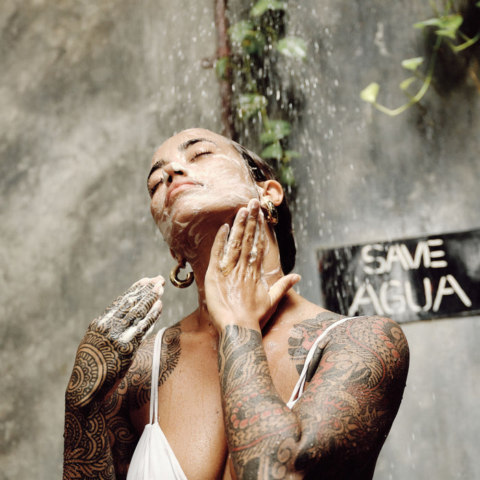 A tattooed person, wearing earrings and a white top, stands under an outdoor shower with eyes closed. They use Thrive Natural Care Face Wash on their face and neck. A sign saying Same Água is visible in the background, with green leaves hanging overhead.
