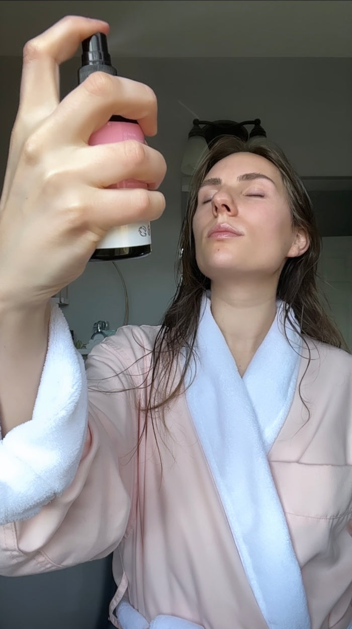 A person in a pink and white bathrobe sprays Beauty by Earths Facial Toner with Witch Hazel & Rose Petal Water on their face. With their eyes closed, they hold the bottle close in a serene bathroom setting, enhancing the rituals tranquility.