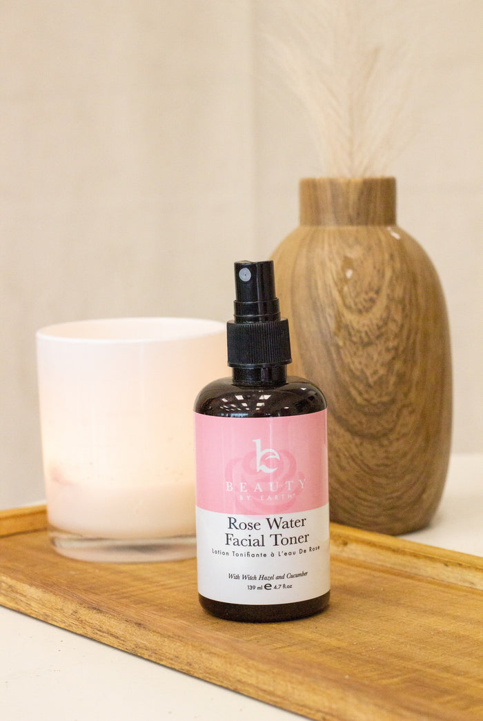 The Facial Toner with Witch Hazel & Rose Petal Water by Beauty by Earth, in a bottle with a pink label, is on a wooden tray. Behind it, a lit white candle and a decorative wooden vase with dried stalks enhance the serene and natural ambiance.