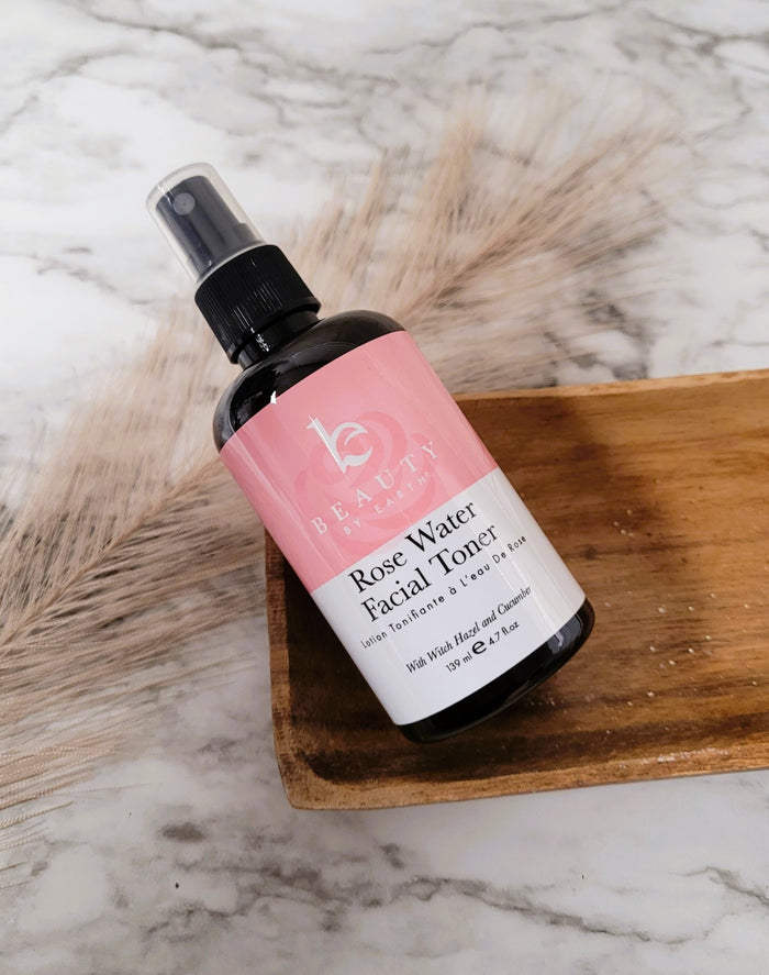 A spray bottle of Beauty by Earths Facial Toner with Witch Hazel & Rose Petal Water, stylishly labeled in pink and white, sits on a wooden tray against a marble background. Accompanied by pampas grass, this serene setup radiates natural charm.