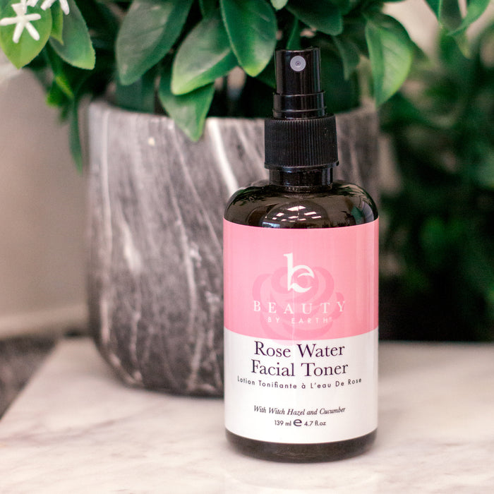 Beauty by Earths Facial Toner with Witch Hazel & Rose Petal Water sits elegantly on marble, its pink and white label popping against a backdrop of lush green leaves from a potted plant.