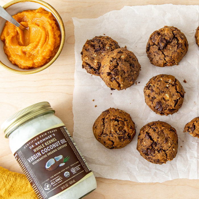 A jar of Dr. Bronners Fair Trade & Organic Whole Kernel Virgin Coconut Oil, a bowl of orange puree with a spoon, and seven chocolate chip cookies sit on parchment paper atop a light wooden surface.