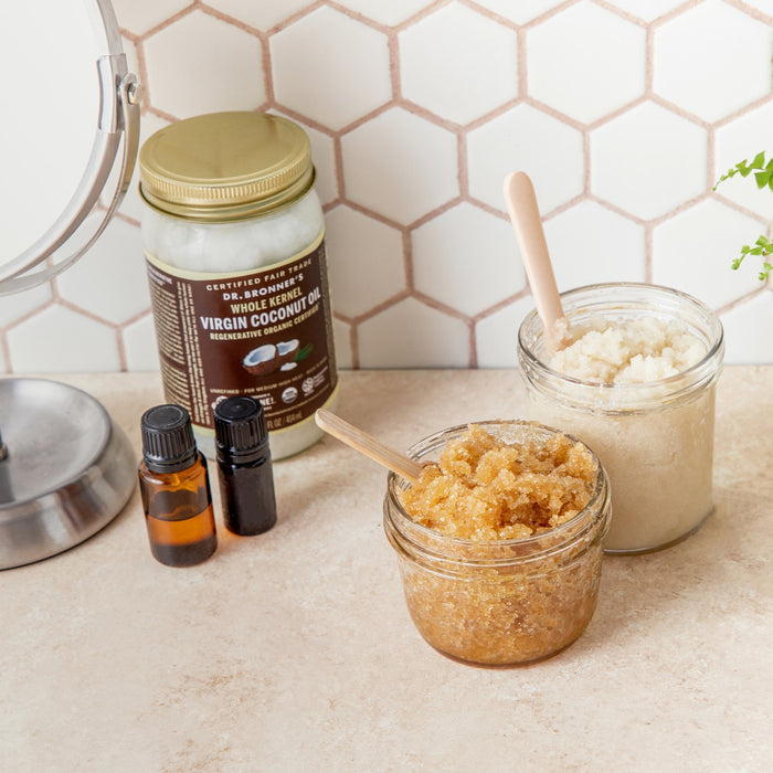 Two jars of homemade sugar scrub with wooden spoons sit on a bathroom counter next to a jar of Dr. Bronners Fair Trade & Organic Whole Kernel Virgin Coconut Oil, two small bottles, and a round mirror against a hexagon tile backsplash.