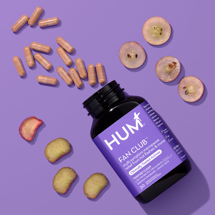 A bottle of HUM Nutritions Fan Club Menopause Relief - For Hot Flashes is open, with capsules spilling onto a purple surface. Slices of grapes and rhubarb are scattered around, highlighting the blend’s natural approach to symptom relief.