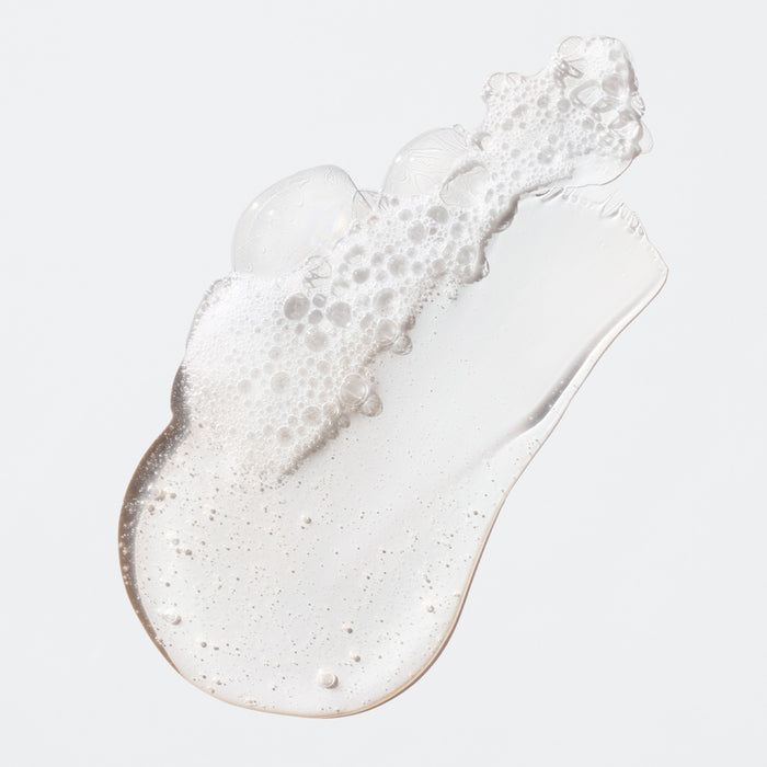 The Fantastic Face Wash Traveler by Ursa Major features a smooth, glossy clear gel with foamy bubbles on a light gray backdrop, capturing the essence of an exfoliating cleanser for soft skin and a refreshing daily routine.