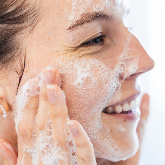 A person with soap lather on their face gently rubs their cheeks, smiling slightly. The image captures a refreshing daily cleanse moment using Ursa Majors Fantastic Face Wash Traveler for a soft skin glow.