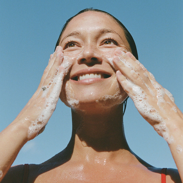 A smiling woman with wet hair uses Ursa Majors Fantastic Face Wash Traveler to cleanse her skin, standing outside under a clear blue sky.