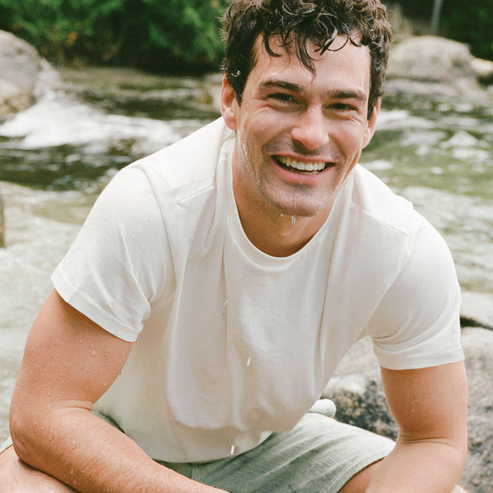 A young man with wet hair and a white t-shirt smiles by a river, enjoying a refreshing cleanse with Ursa Majors Fantastic Face Wash Traveler, surrounded by rocks and greenery.