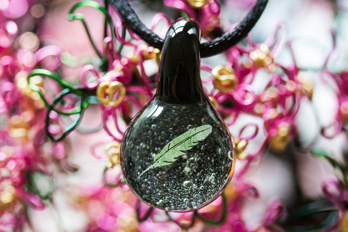 Close-up of a dark glass pendant with a green holographic feather inside hanging from a black cord