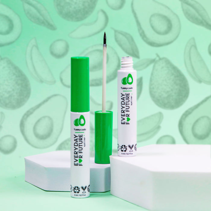 Two Everyday For Future Avocado Felt Tip Eyeliners rest on white geometric platforms, with one tube open to reveal a brush applicator. Set against a green backdrop with avocado illustrations, these eyeliners are made from vegan and cruelty-free ingredients.