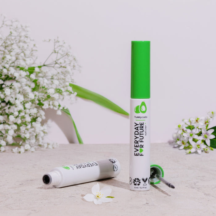 Two green-top Felt Tip Avocado Eyeliner tubes by Everyday For Future, one open and one closed, lie on a stone surface. Surrounded by white flowers and green leaves, the scene suggests freshness and highlights the vegan, cruelty-free nature of these products featuring castor oil.