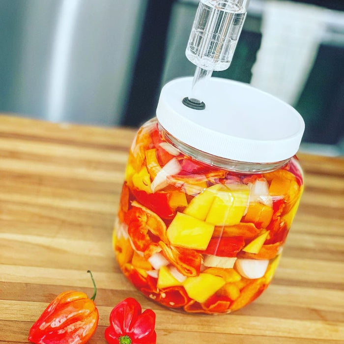 On a wooden countertop sits the FarmSteady Fermented Hot Sauce Kit, containing a glass jar of sliced orange and red peppers, onions, and mango. The lid has an attached fermentation airlock. Two whole habanero peppers rest nearby.